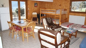 Ein gemütliches Zimmer mit einem Holzstockbett, einem Esstisch mit Stühlen, einem kleinen Tisch mit Büchern und Blick ins Freie durch ein Fenster.