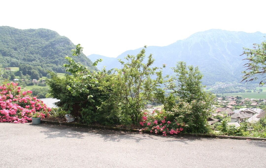 Uma estrada com uma fileira de flores cor-de-rosa e arbustos verdes, que leva a uma aldeia com casas e montanhas ao fundo.