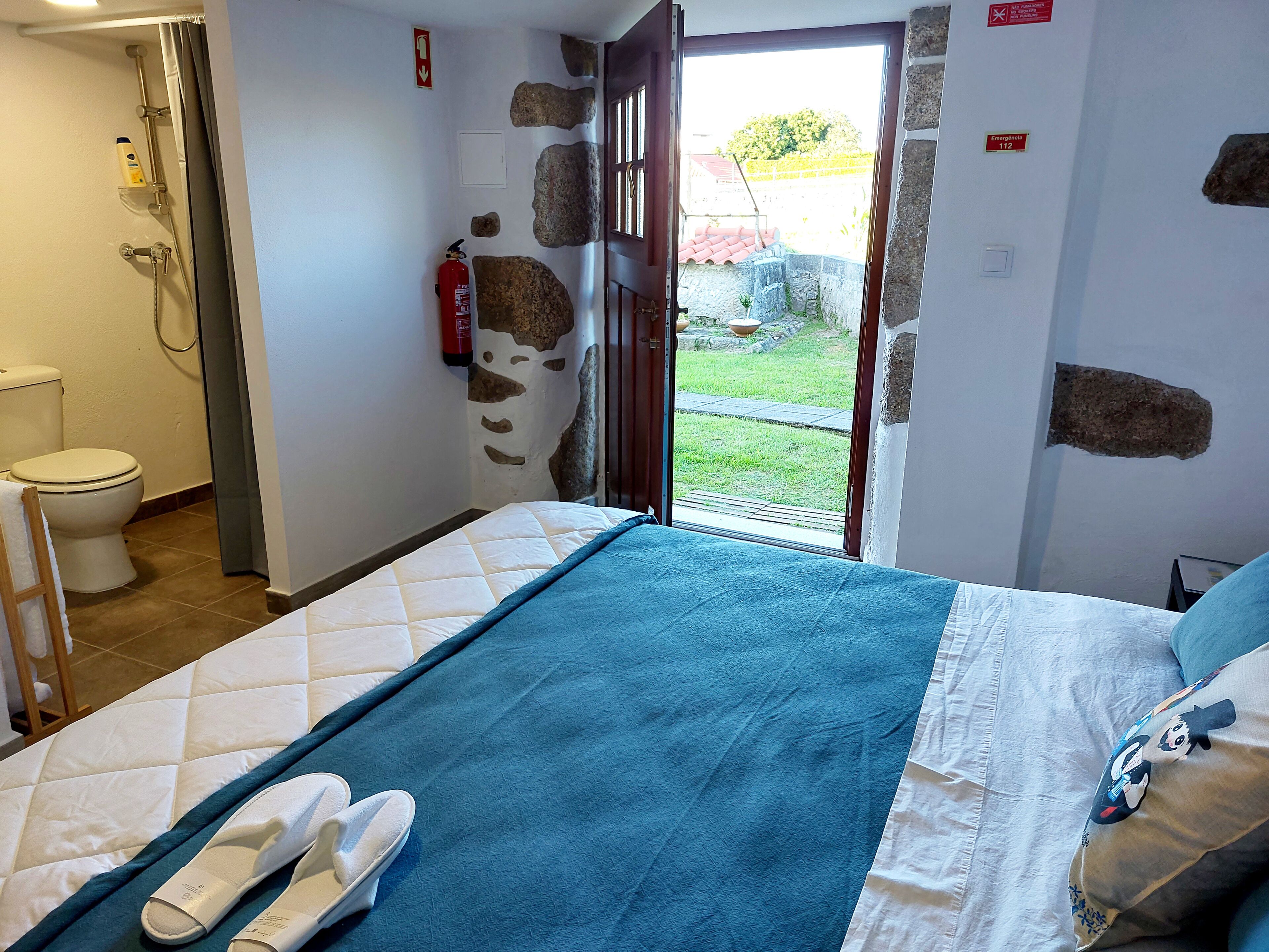 Habitación de hotel con cama, baño con ducha, inodoro y extintor.