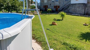Una piscina con bordo bianco, un edificio in pietra con tetto in tegole rosse e un prato con un piccolo albero e una fioriera.