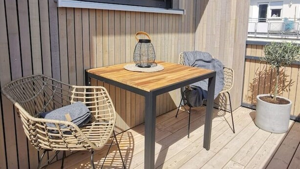 A wooden deck with a table and two chairs, a potted plant, and a window with blinds.