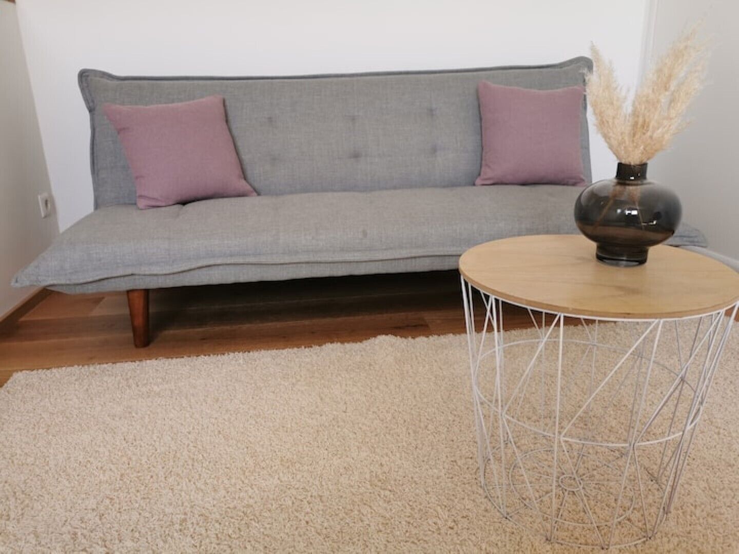 En grå sofa med to puder, et rundt træsofabord med metalstel og en beige, luvrigt måtte.