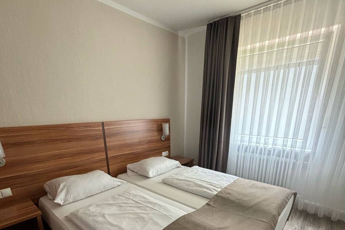 Un lit double avec une tête de lit en bois, deux tables de chevet et une grande fenêtre ornée de rideaux légers.