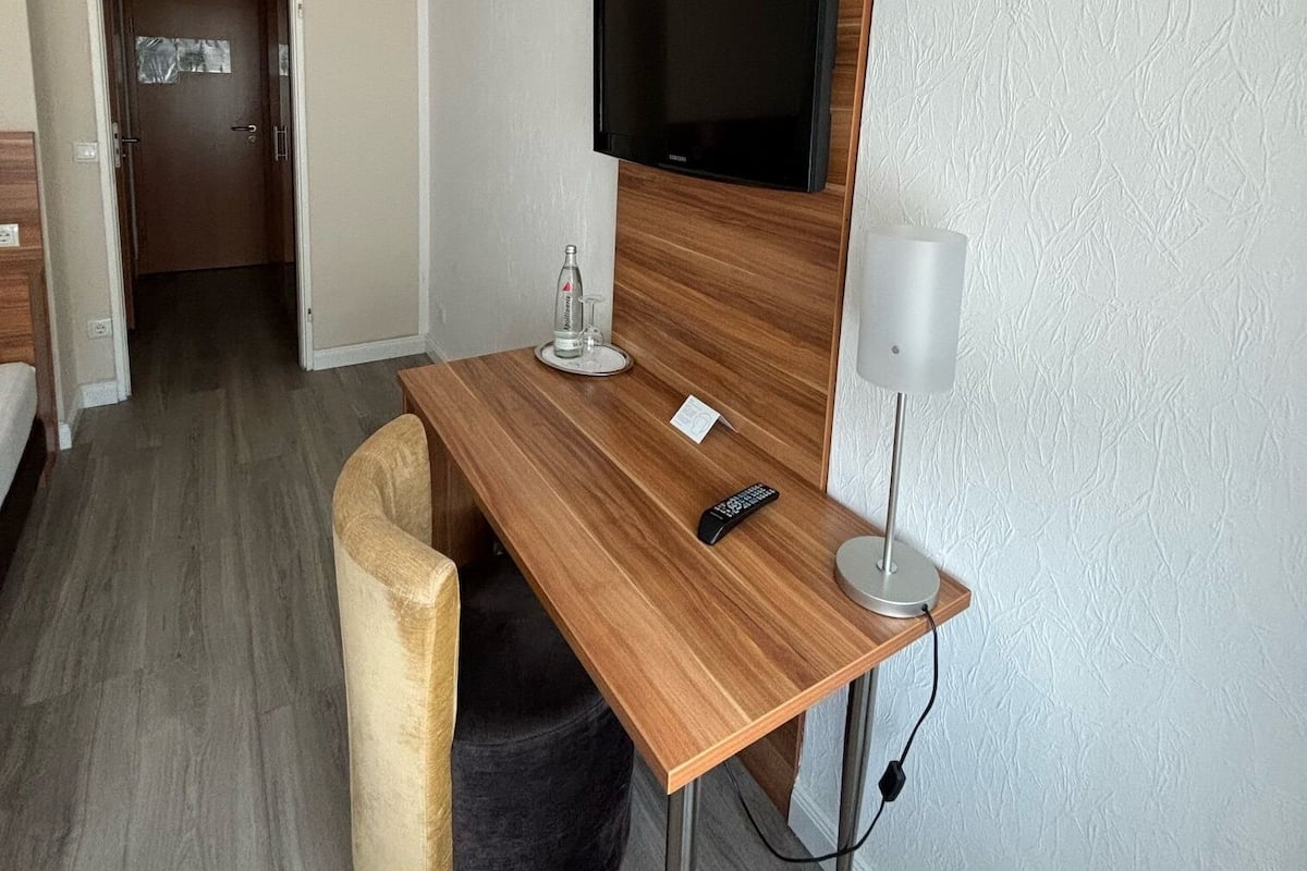 Une chambre d’hôtel moderne avec un bureau en bois, un téléviseur à écran plat fixé au mur, un petit pouf rond et une table de chevet avec une lampe.