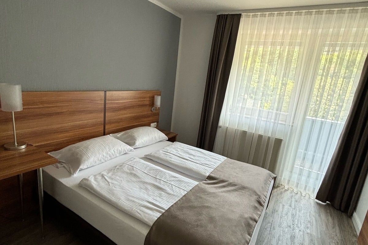 Une chambre d’hôtel avec une tête de lit en bois, un grand lit avec du linge de lit blanc, une table de chevet avec une lampe et une fenêtre ornée de rideaux légers.