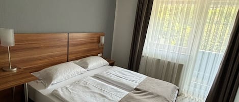 Una habitación de hotel con cabecera de madera, una cama grande con ropa blanca, una mesita de noche con lámpara y una ventana con cortinas translúcidas.