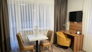 Hotelkamer met een eettafel, twee stoelen, een klein bureau met stoel, een aan de muur bevestigde televisie en een raam met dunne gordijnen.