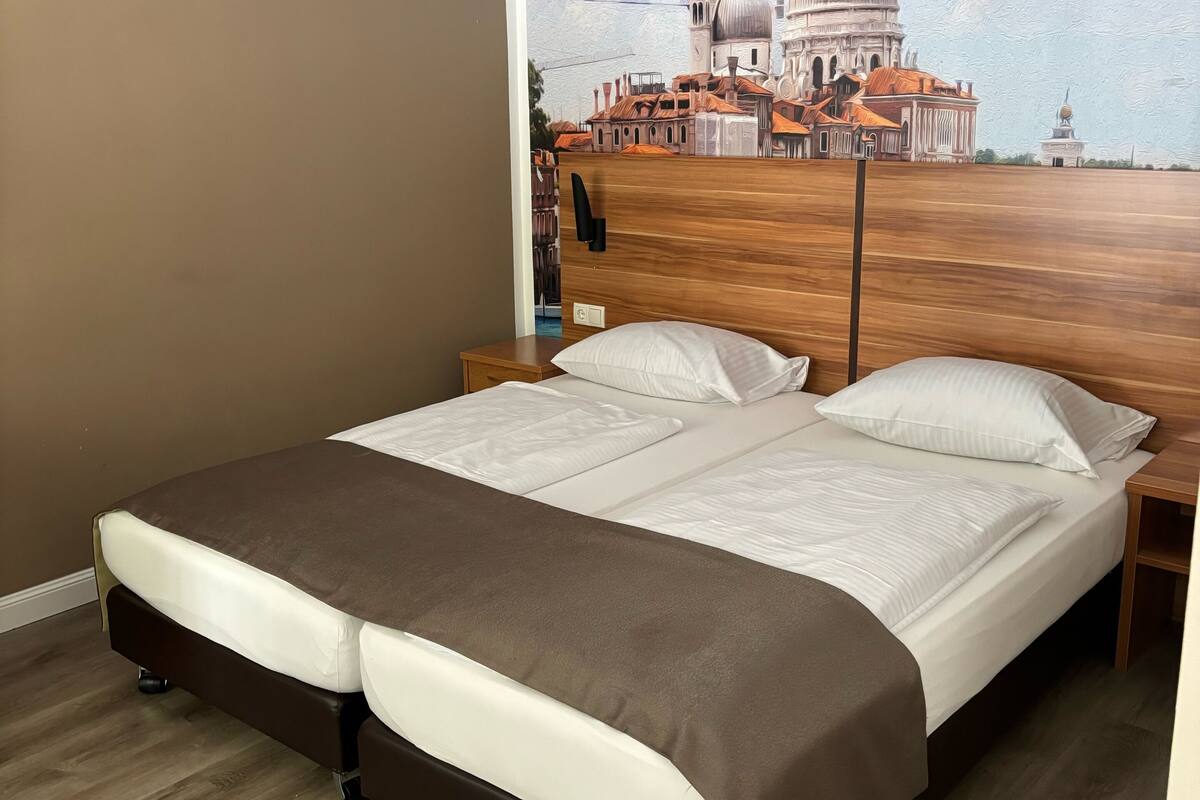 Un lit double avec une tête de lit marron et du linge de lit blanc, dans une chambre dont un des murs est orné d’une grande fresque représentant un paysage urbain.