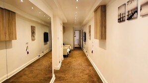Un pasillo con piso alfombrado, paredes blancas y paneles de madera. Se observan cuadros enmarcados en las paredes y una puerta al final.
