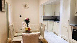 Un comedor con mesa de vidrio y sillas acolchadas, un florero con flores y un reloj en la pared. Junto a él, una cocina con gabinetes blancos y encimera negra.