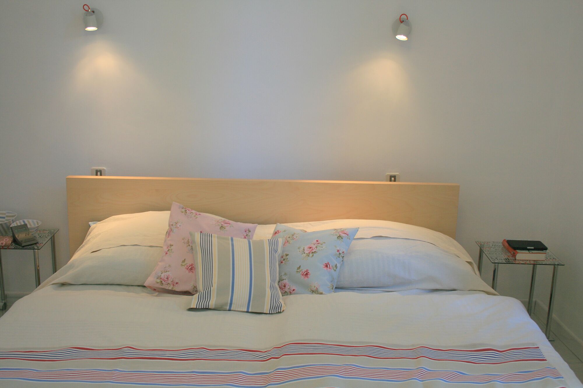 A neatly made bed with floral and striped pillows, a plain headboard, and bedside tables with a lamp and a book.
