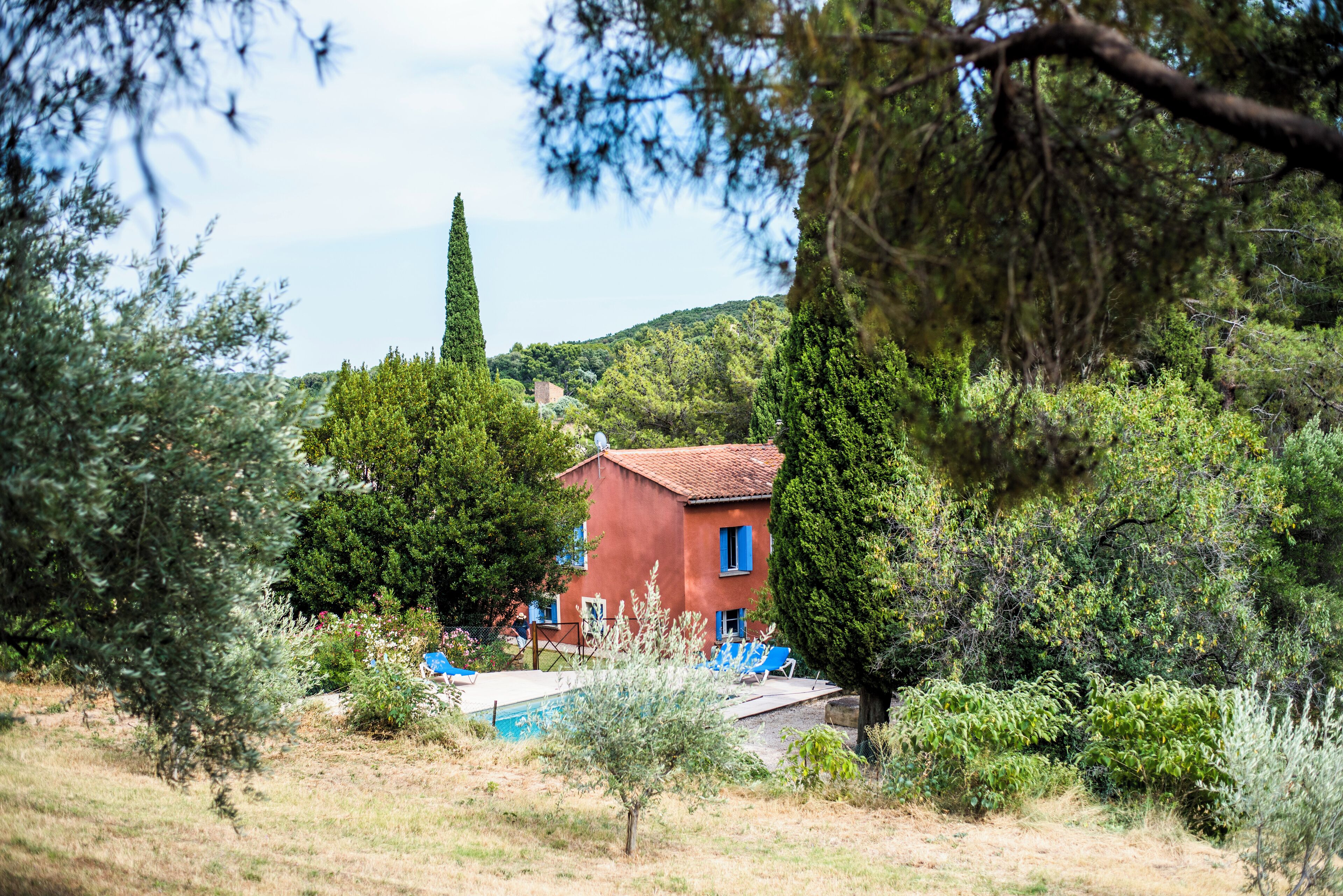 Un bâtiment rouge aux volets bleus, entouré de verdure et bordé d’une piscine.