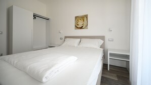 Ein ordentlich bezogenes Bett mit weißen Laken, ein weißer Nachttisch und ein an der Wand hängendes Bild von einer Blume.