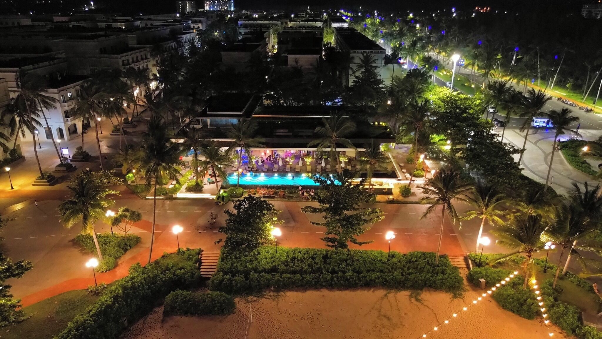 Une vue nocturne d’un hôtel avec une piscine, des palmiers et une rue bien éclairée.