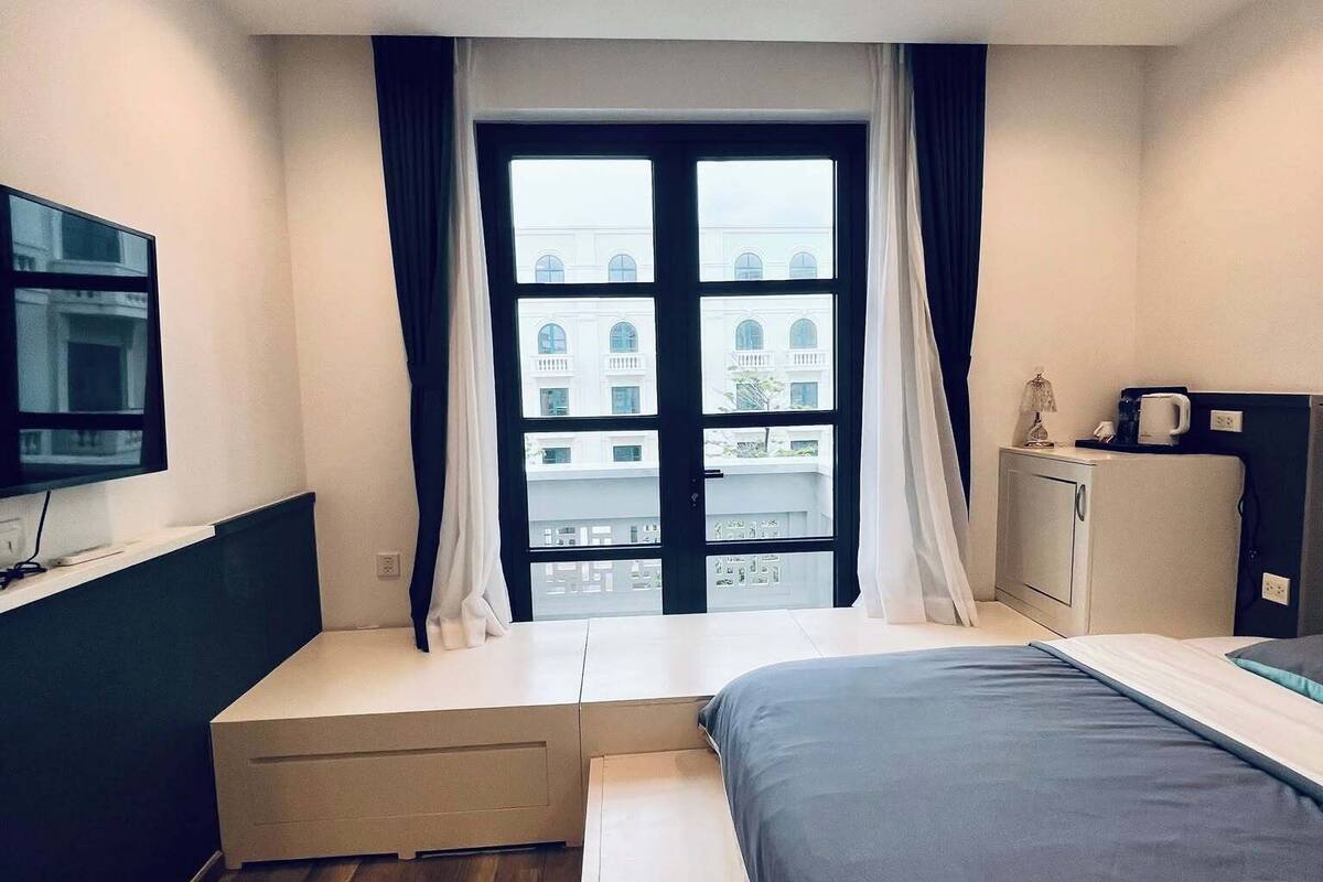 A modern hotel room with a bed, a desk, a flat-screen TV, a window with curtains, and a view of buildings outside.