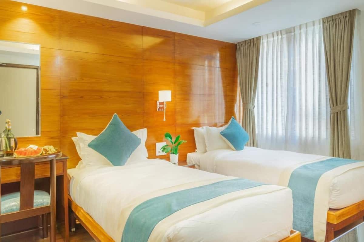 A hotel room with two beds, wooden paneling, a nightstand with a lamp, a plant, and a window with curtains.