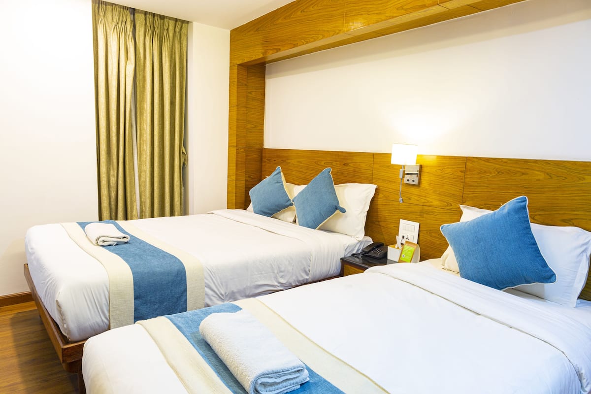 A hotel room with two beds, wooden headboard, white linens, blue pillows, a nightstand with a phone and a lamp, and a curtain on the window.