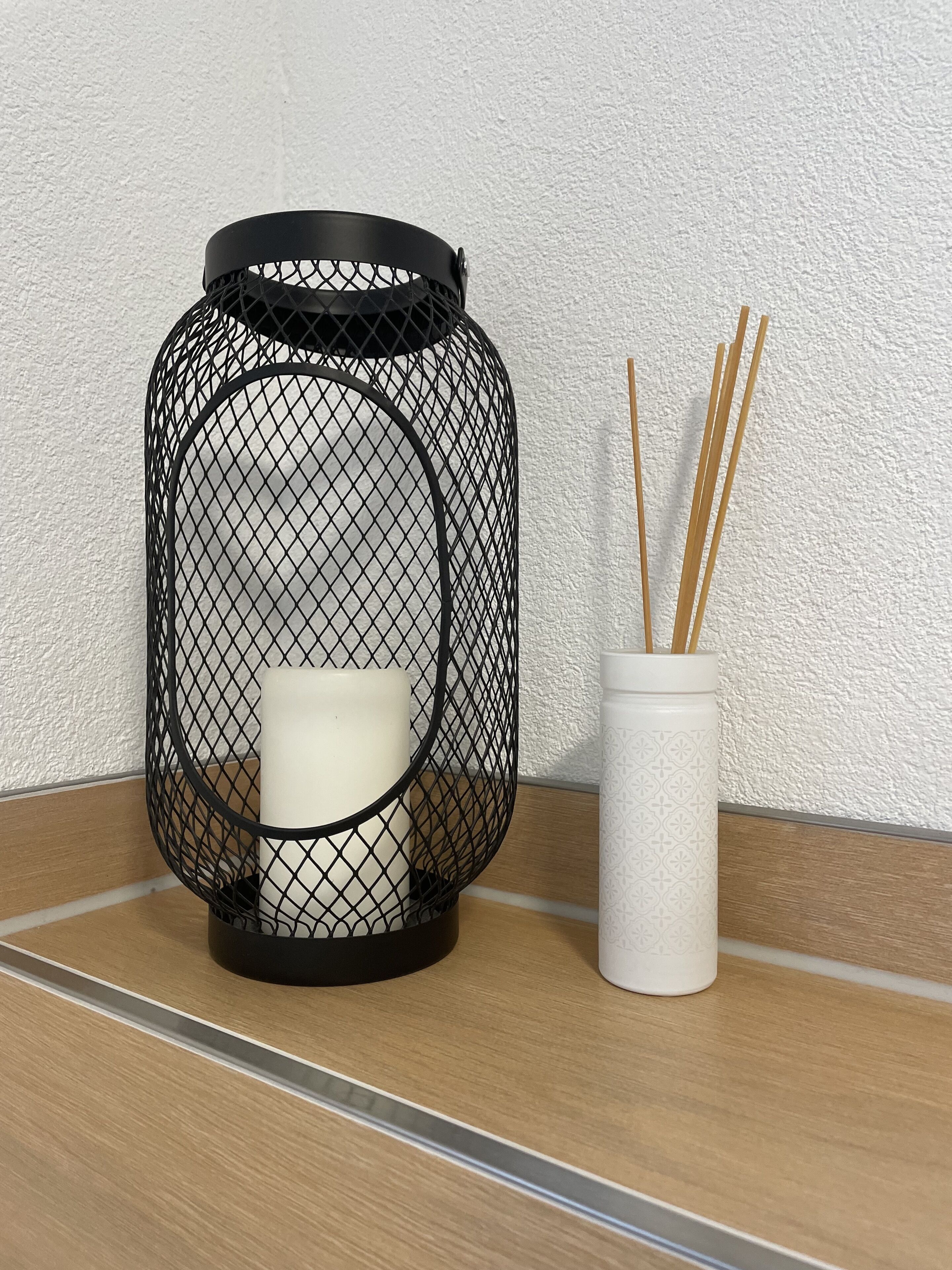 Une lanterne noire à armature métallique, avec une bougie blanche à l’intérieur, posée sur une surface en bois, à côté d’un vase blanc contenant des tiges de roseau.