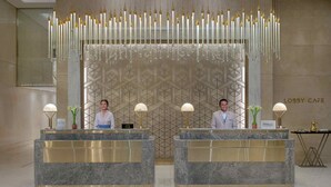 Zona de recepción de hotel con dos mostradores, cada uno atendido por un recepcionista. Hay luces decorativas colgantes sobre los mostradores y un papel tapiz con motivos en la pared de fondo.