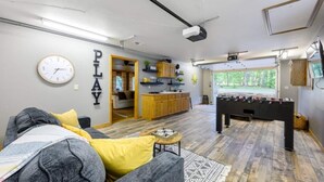 Een woonkamer met een bank, een tafelvoetbaltafel en een keuken, zichtbaar door een open deur.
