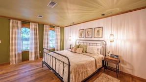 Een slaapkamer met een metalen bedframe, een houten vloer, een raam met gordijnen en een nachtkastje met een lamp.