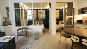 Habitación de hotel moderna con una cama grande, una mesa de comedor, dos sillas, una mesita redonda pequeña, un sofá, una cafetera y un televisor.