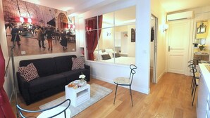 Una sala de estar moderna con un sofá negro, una mesa de centro blanca y un espejo grande que refleja una cama y una ventana.