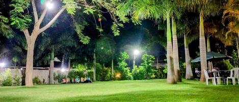 A well-lit outdoor area with a grassy lawn, trees, and a seating area with tables and chairs.