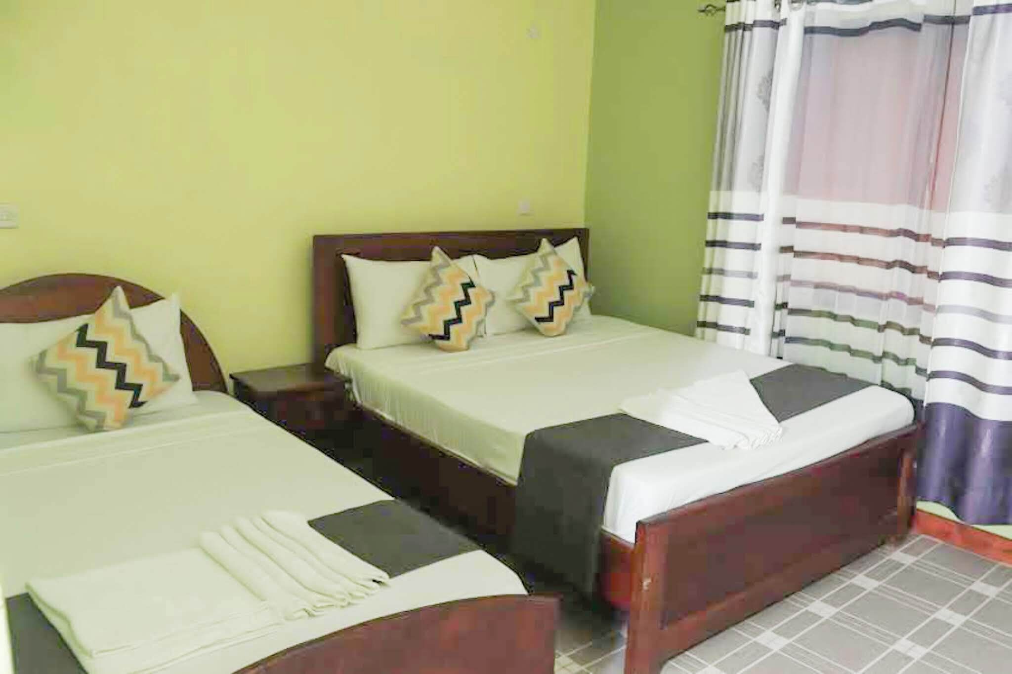 A hotel room with two single beds, each with a wooden headboard, white bedding, and decorative pillows. There is a nightstand between the beds, and a window with a striped curtain.