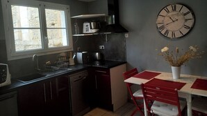 A compact kitchen with a black stove, a microwave, and a sink. There is a dining table with red chairs and a vase of flowers. A large clock is mounted on the wall. - House in historic center of Caen! (Caen)