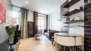 Ein modernes Wohnzimmer mit einer Couch, einem Couchtisch und einem Essbereich mit zwei Stühlen. Ein Bücherregal mit Büchern und dekorativen Gegenständen ist ebenfalls vorhanden.
