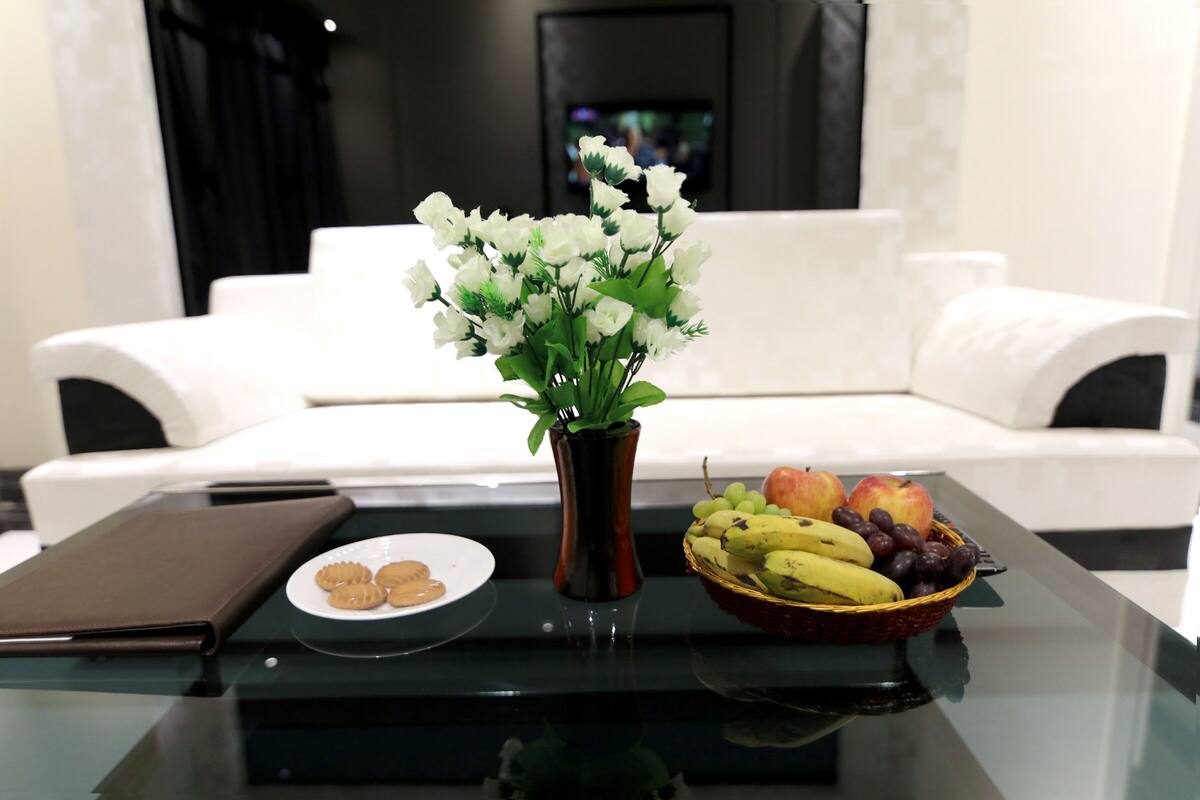 Een moderne woonkamer met een witte bank, een glazen bijzettafeltje met een vaas witte bloemen, een bord koekjes en een fruitmand.