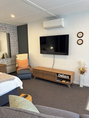 Ein modernes Hotelzimmer mit einem Flachbildfernseher, einem Holzkonsolentisch mit einem Schild mit der Aufschrift 'Believe', einer Ziegelwand und einem großen Spiegel.
