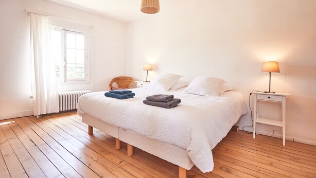 A bedroom with a large bed, two bedside tables with lamps, a radiator, and a window with curtains. - The Grand Sambam (Fourdrain)