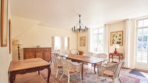 A dining room with a wooden table and wicker chairs, a chandelier, and a large window with curtains. - The Grand Sambam (Fourdrain)