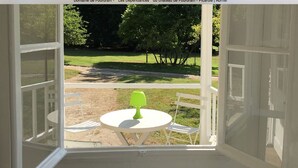 An open balcony with a white table and chairs, a green table lamp, and a view of a grassy area with trees. - The Grand Sambam (Fourdrain)