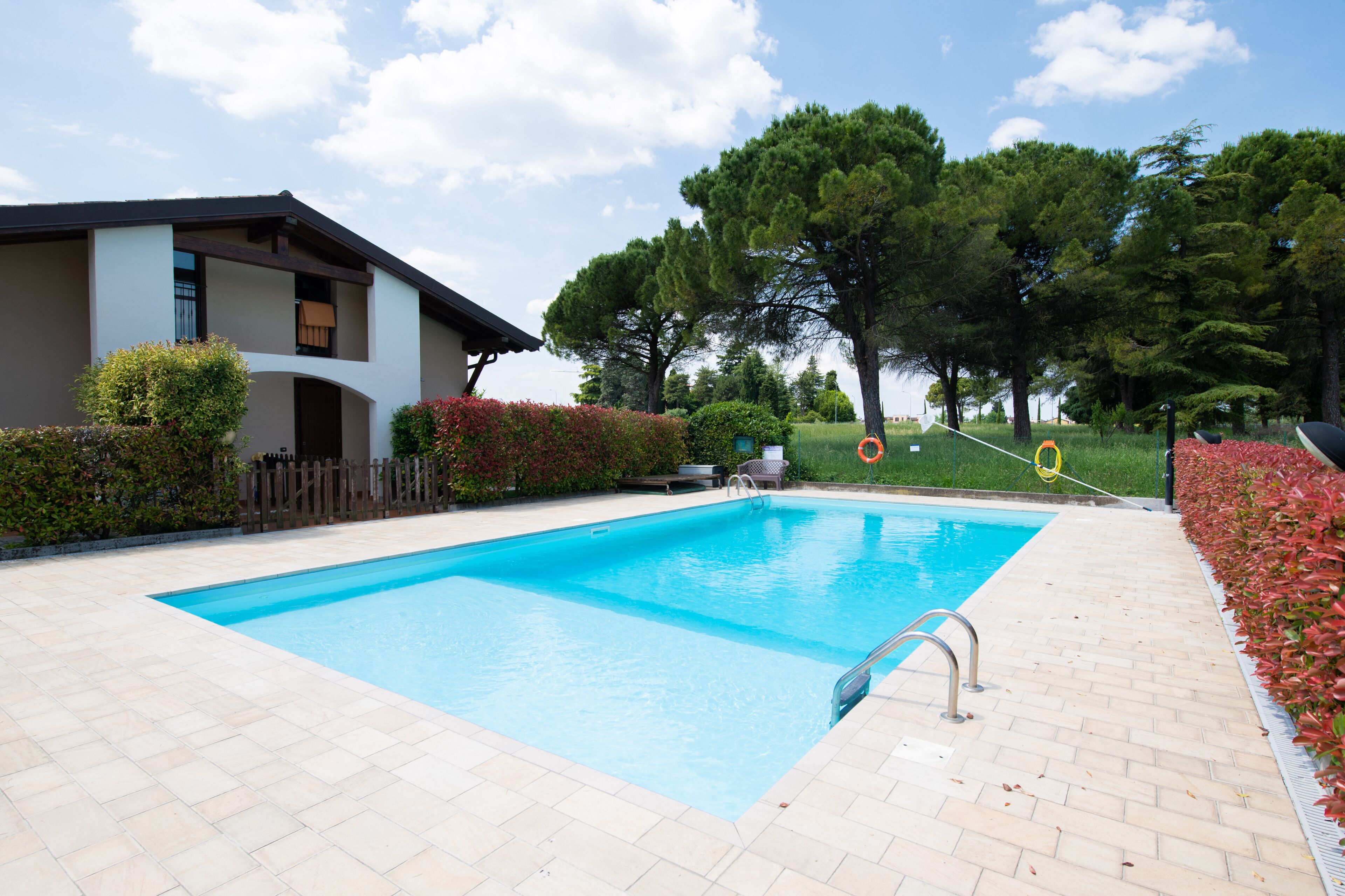 A rectangular swimming pool with a tiled deck, surrounded by greenery and a building with a balcony.