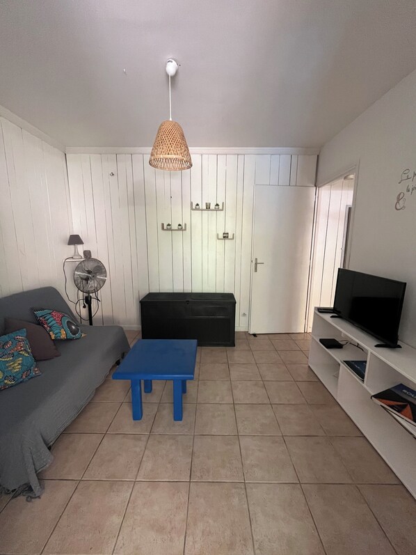 A living room with a sofa, a blue table, a TV on a stand, a fan, and a door with a shelf above it. - Gites Les Vanilles: lush tropical garden, pool 800 meters from the beach (Deshaies)