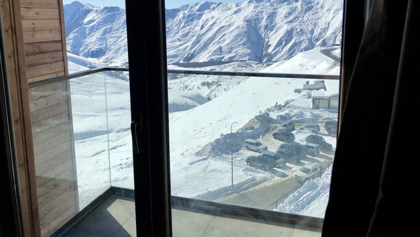 A view from a window with a metal railing, overlooking a snowy mountainous landscape with buildings and vehicles. - 2-room apartment NFT 452 New Gudauri (Gudauri)