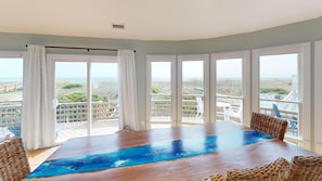 Una amplia mesa de madera con incrustación azul, rodeada de sillas de mimbre, en una habitación con grandes ventanales que dan a la playa y al océano.