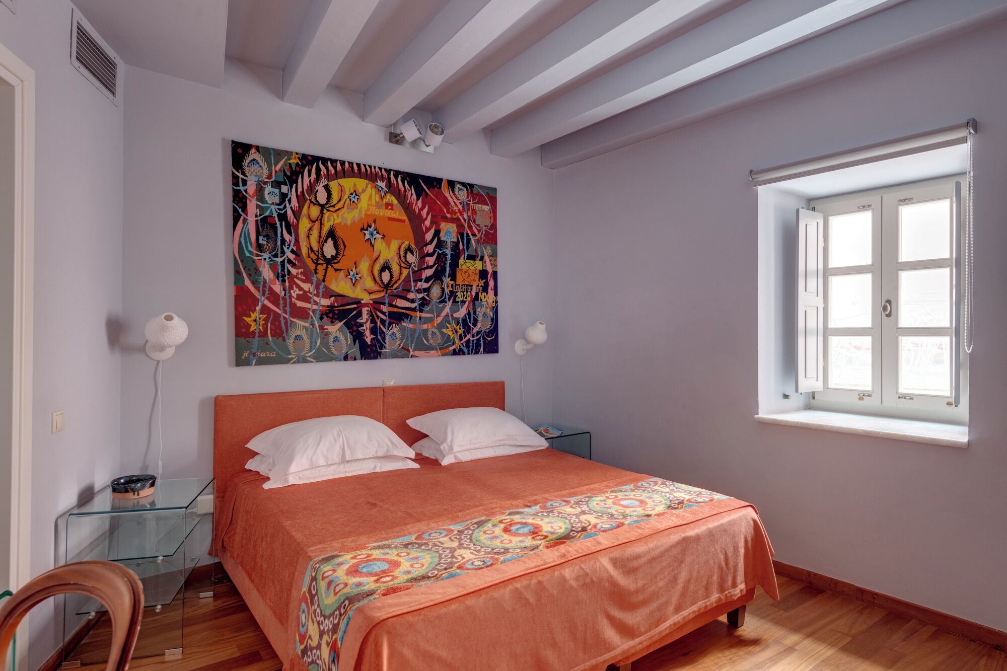 A bedroom with a bed, a colorful painting, a window, and a bedside table.