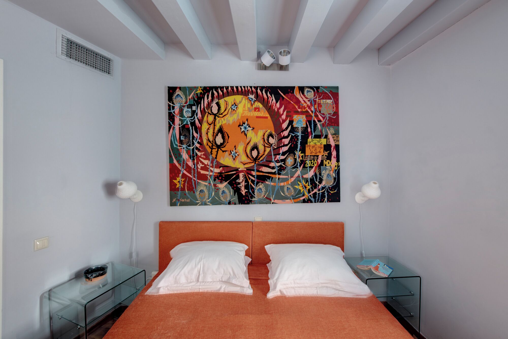Un dormitorio con un cuadro abstracto grande, una mesita de noche de cristal con un libro y una mesita auxiliar de cristal con una lámpara.