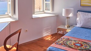 Una habitación con cama, una silla, una mesa y una lámpara. Hay ventanas con vistas al mar y a un edificio.