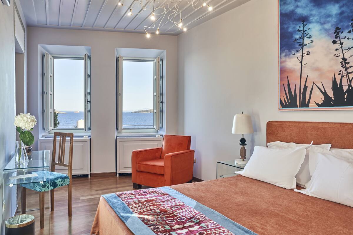 Ein Schlafzimmer mit einem großen Bett, zwei Fenstern mit Meerblick, einem Sessel, einem Tisch mit Blumenvase und einem Bild an der Wand.