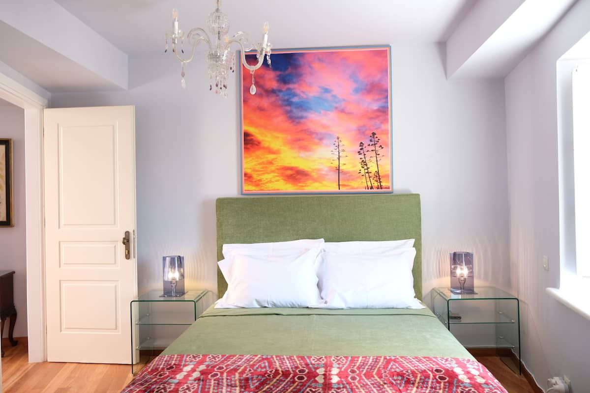 Ein Schlafzimmer mit einem grünen Kopfteil, weißer Bettwäsche, einem farbenfrohen Sonnenuntergangsbild und zwei Nachttischen mit Lampen.