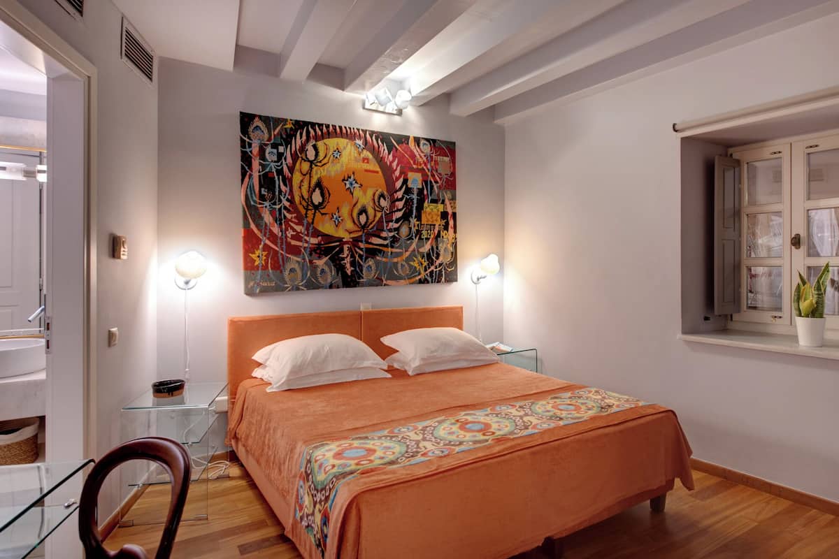 Ein Schlafzimmer mit einem großen Bett, zwei Nachttischlampen, einem Fenster mit Vorhängen und einem farbenfrohen, abstrakten Gemälde an der Wand.
