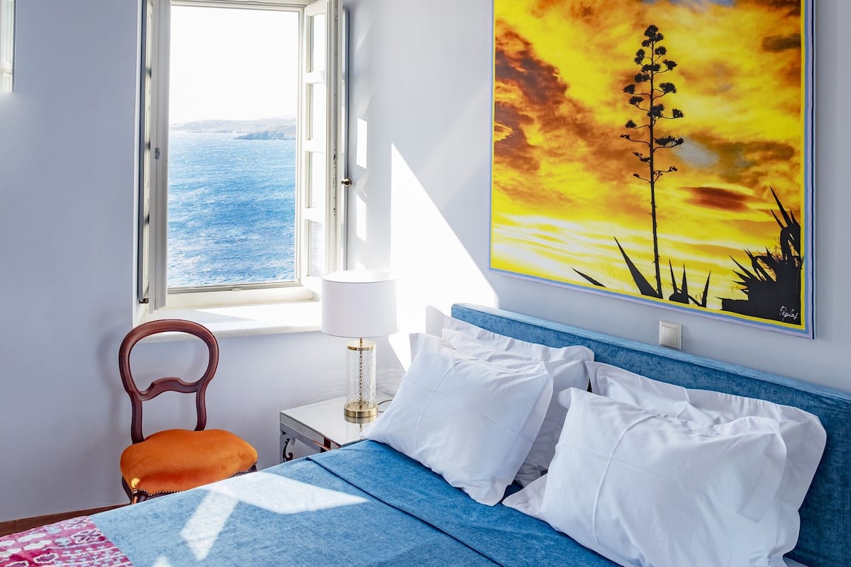 Ein Schlafzimmer mit einem Bett, einem Fenster mit Meerblick, einem Sonnenuntergangsbild und einer Chandelier.