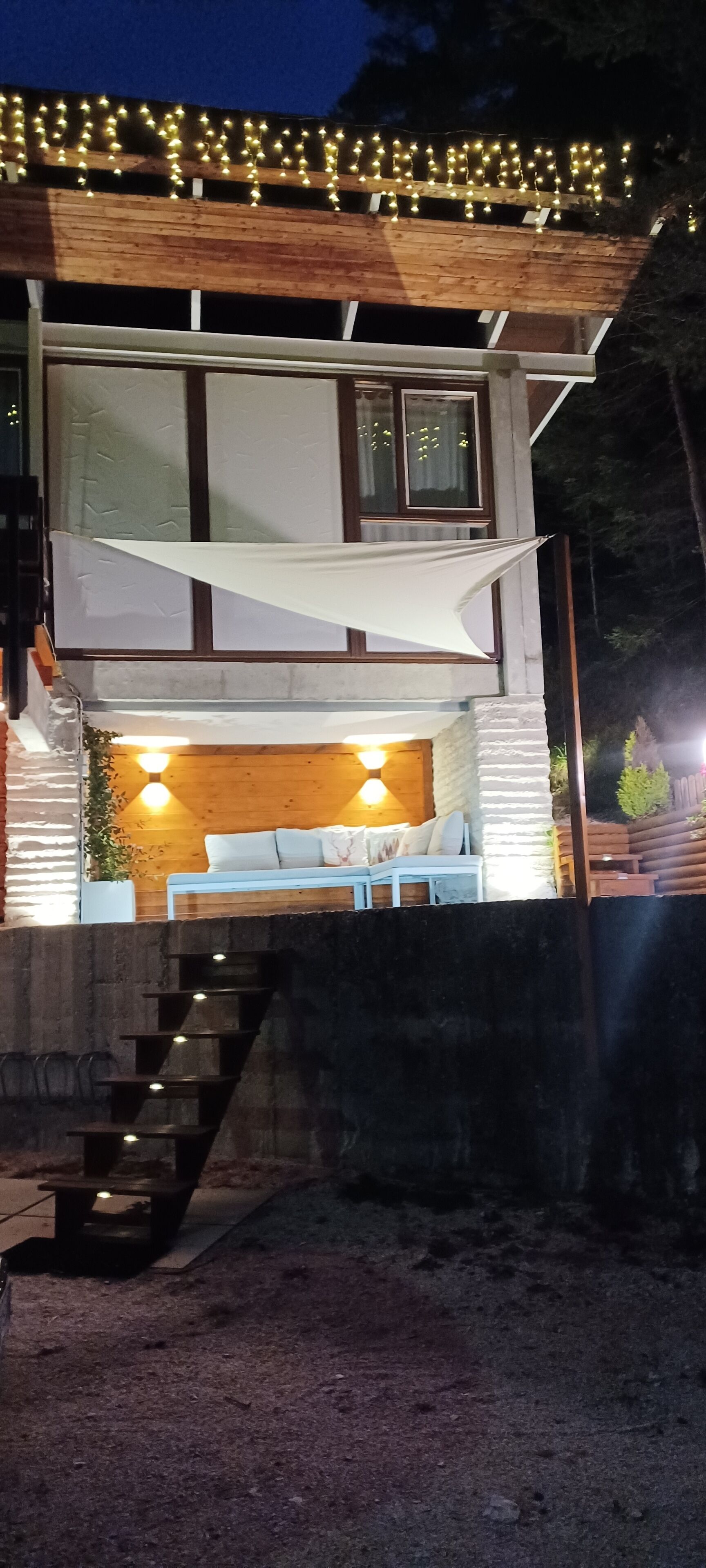 Ein modernes Haus mit einer Holzterrasse, einem weißen Markisenvorbau und einer Sitzgruppe mit Kissen.