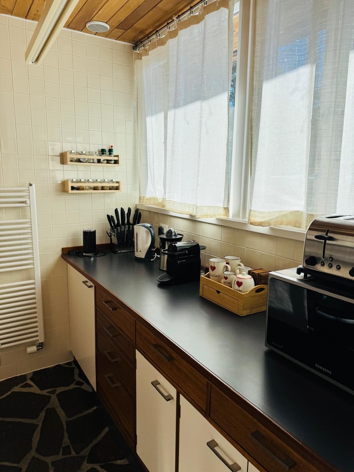 A kitchen with a built-in oven, a coffee maker, and a kettle on the counter. There are wooden cabinets and a window with curtains.