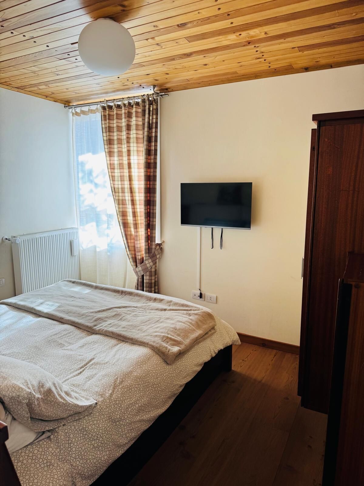 A bedroom with a wooden ceiling, a bed with a beige blanket, a television on the wall, and a window with plaid curtains.